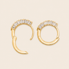 A pair of gold crescent hoop earrings with a row of clear baguette-cut stones along the top curve, displayed on a light beige background.
