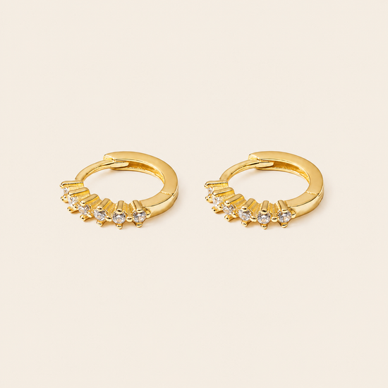A pair of gold huggie hoop earrings with small clear gemstones set along the front, displayed on a light beige background.