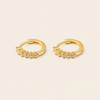 A pair of gold huggie hoop earrings with small clear gemstones set along the front, displayed on a light beige background.