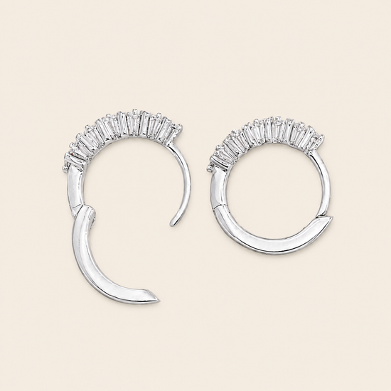 A pair of silver crescent-shaped hoop earrings with a row of clear baguette stones across the top, displayed on a light beige background.