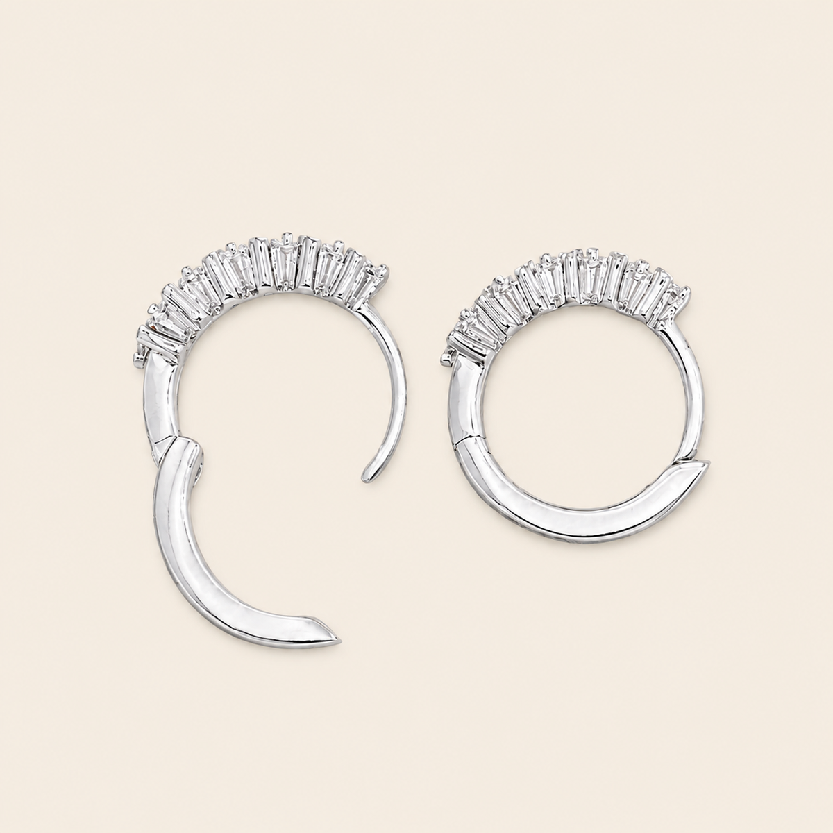 A pair of silver crescent-shaped hoop earrings with a row of clear baguette stones across the top, displayed on a light beige background.