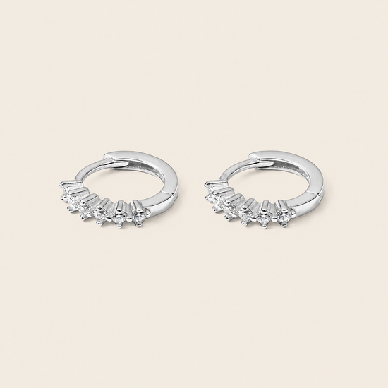 A pair of silver huggie hoop earrings with small clear gemstones set along the front, displayed on a light beige background.