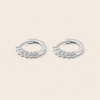 A pair of silver huggie hoop earrings with small clear gemstones set along the front, displayed on a light beige background.