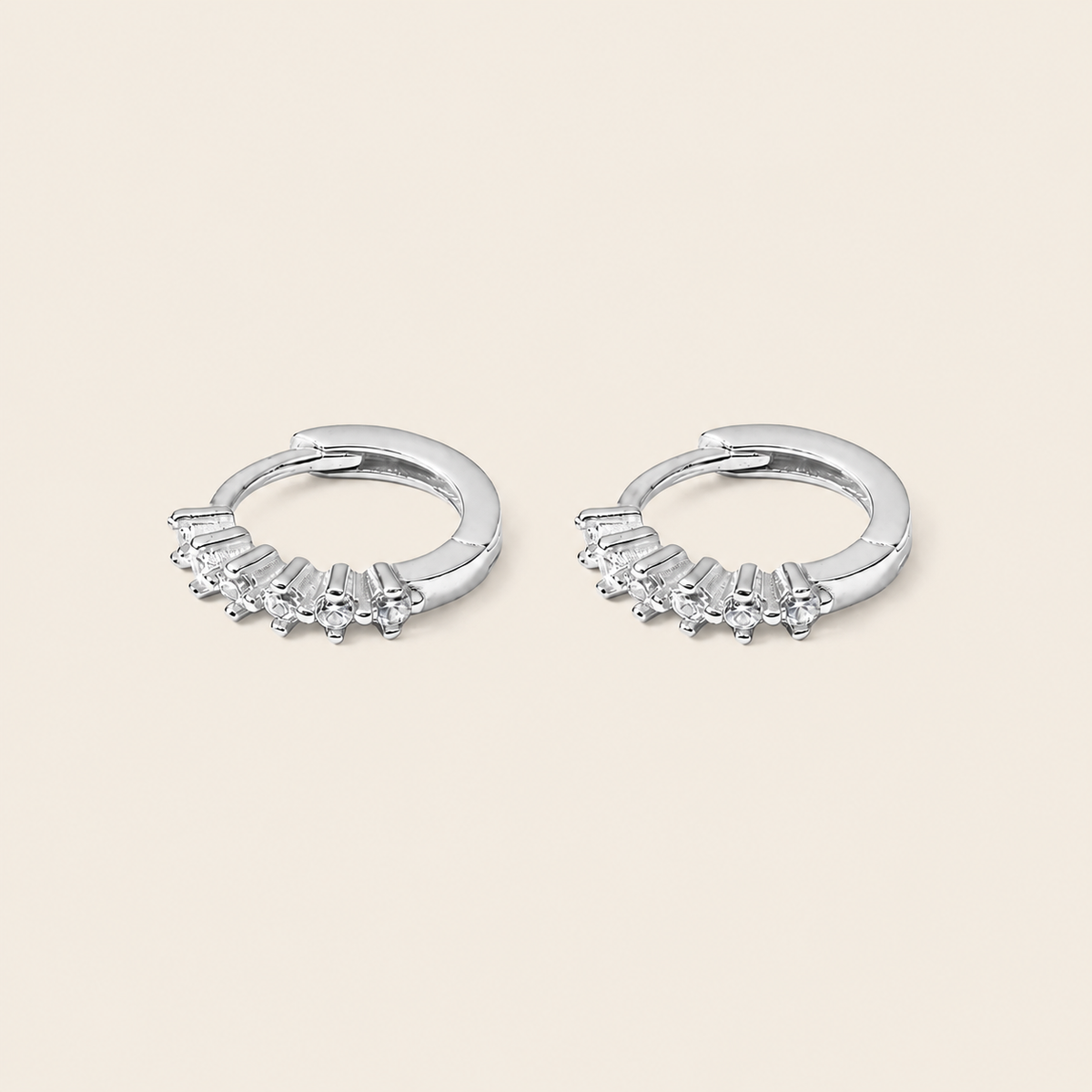 A pair of silver huggie hoop earrings with small clear gemstones set along the front, displayed on a light beige background.