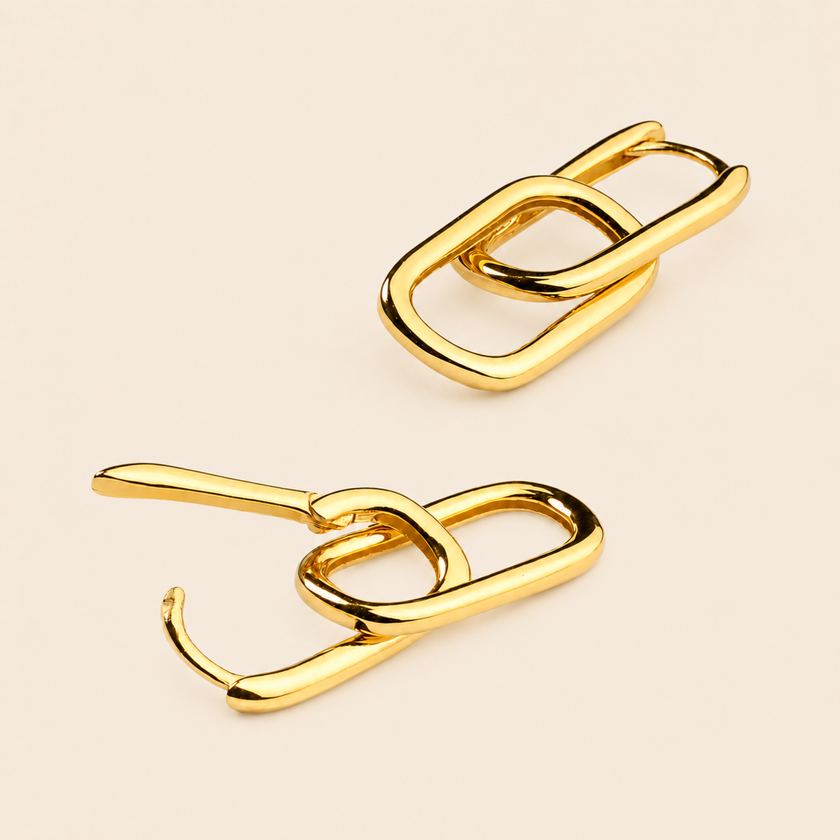 Two gold-toned abstract chain-link earrings with a polished metallic finish, displayed on a light beige background.