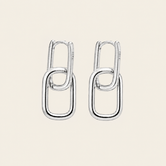 A pair of polished silver drop earrings with interlocking oval link designs, displayed on a solid light beige background.