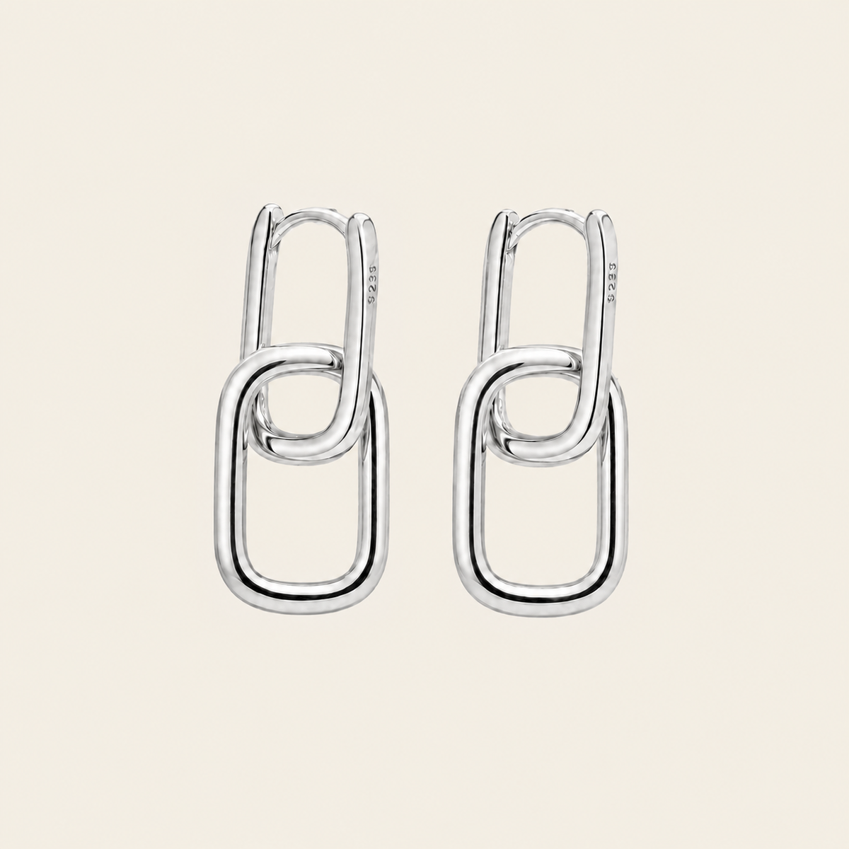 A pair of polished silver drop earrings with interlocking oval link designs, displayed on a solid light beige background.