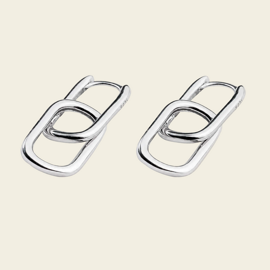 A pair of polished silver-toned interlocking oval link earrings on a solid light beige background.