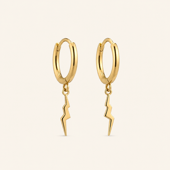 A pair of gold huggie hoop earrings with a dangling lightning bolt charm, shown on a light cream background.
