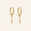 A pair of gold huggie hoop earrings with a dangling lightning bolt charm, shown on a light cream background.