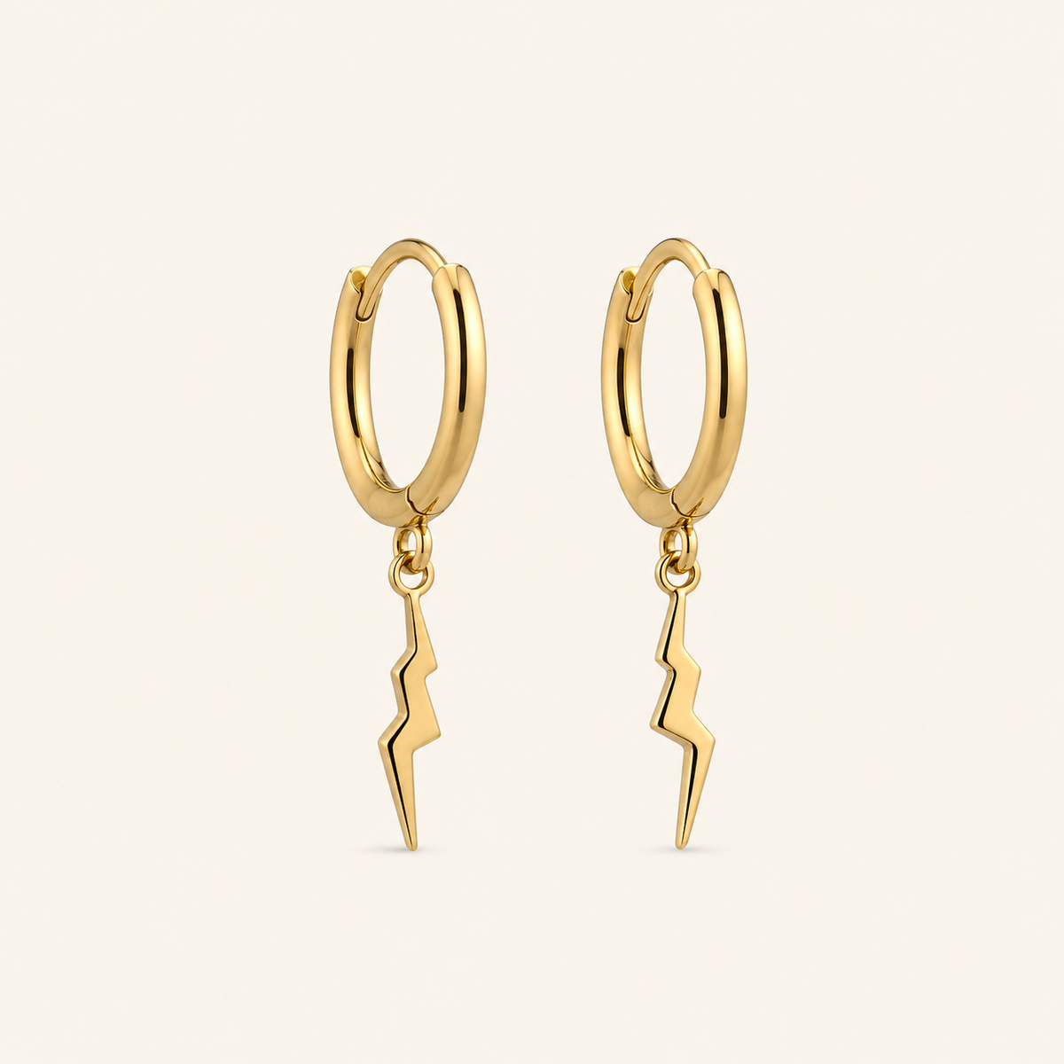 A pair of gold huggie hoop earrings with a dangling lightning bolt charm, shown on a light cream background.