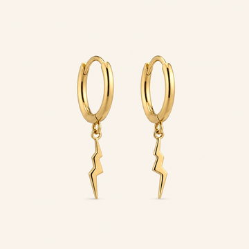 A pair of gold huggie hoop earrings with a dangling lightning bolt charm, shown on a light cream background.