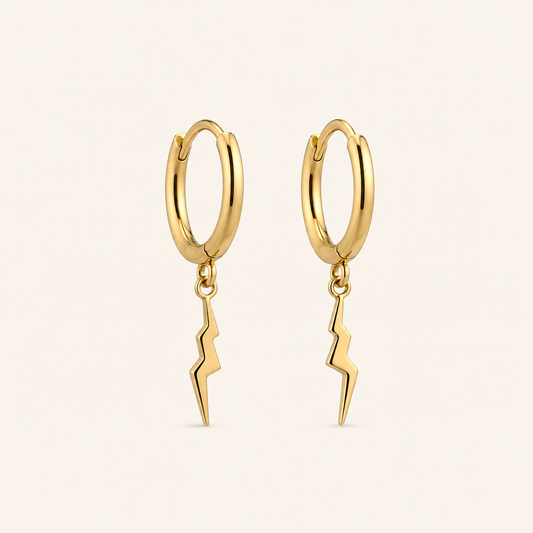 A pair of gold huggie hoop earrings with a dangling lightning bolt charm, shown on a light cream background.
