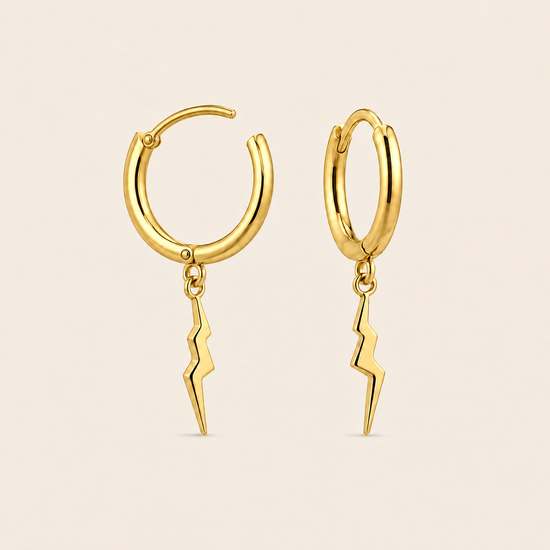 A pair of gold huggie hoop earrings with dangling lightning bolt charms, shown on a solid pale beige background.