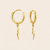 A pair of gold huggie hoop earrings with dangling lightning bolt charms, shown on a solid pale beige background.
