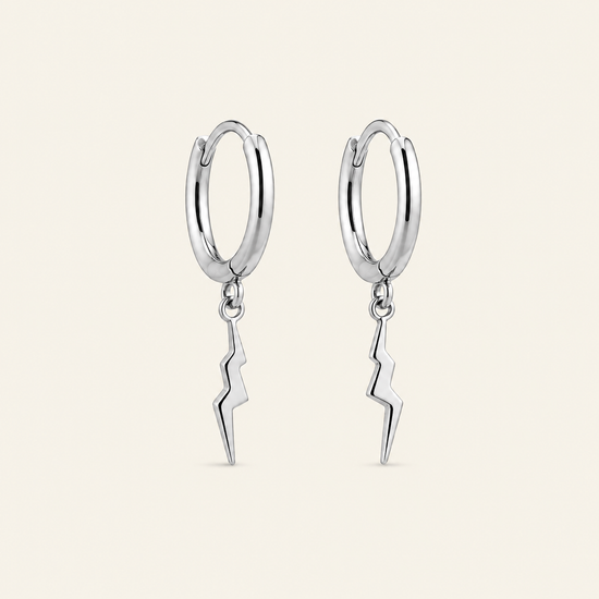 A pair of polished silver hoop earrings with dangling lightning bolt charms, shown against a solid light beige background.