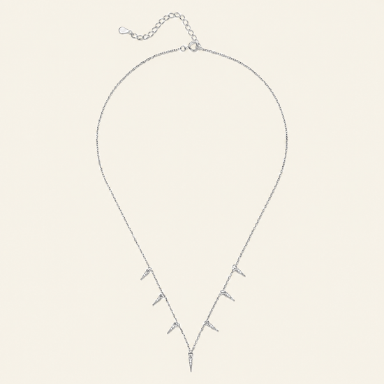 A delicate silver necklace with a V-shaped pendant featuring multiple small spike-like charms, displayed on a solid cream-colored background.