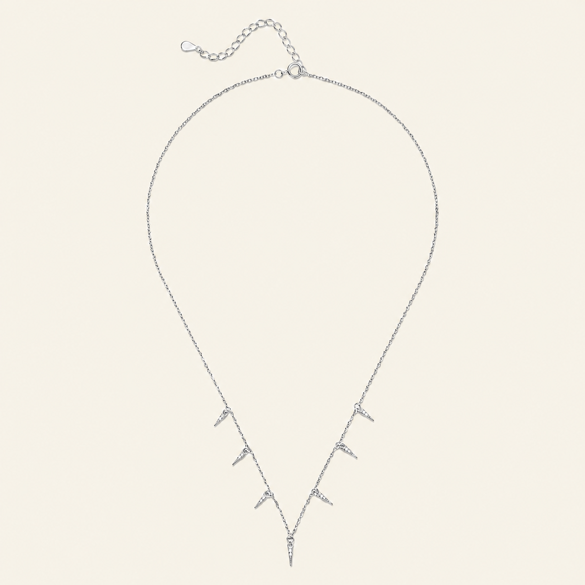 A delicate silver necklace with a V-shaped pendant featuring multiple small spike-like charms, displayed on a solid cream-colored background.