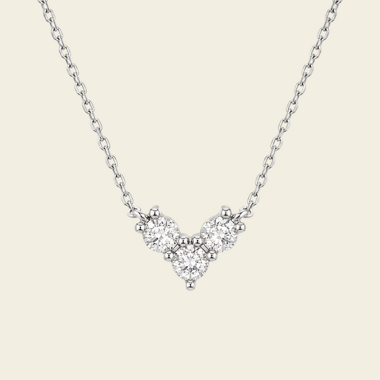 A delicate silver chain necklace featuring a small V-shaped cluster of clear gemstones in a silver setting, displayed against a solid light beige background.