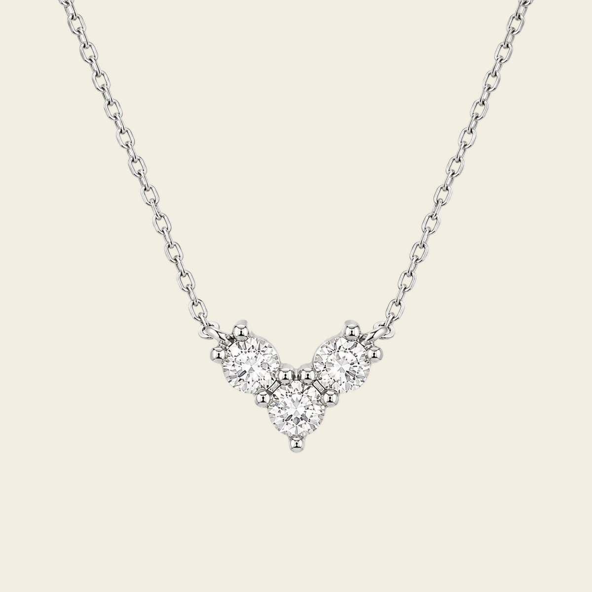 A delicate silver chain necklace featuring a small V-shaped cluster of clear gemstones in a silver setting, displayed against a solid light beige background.