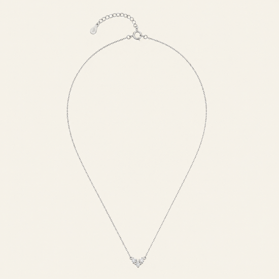 A delicate silver chain necklace with a small heart-shaped pendant set with clear stones, displayed on a light cream background.
