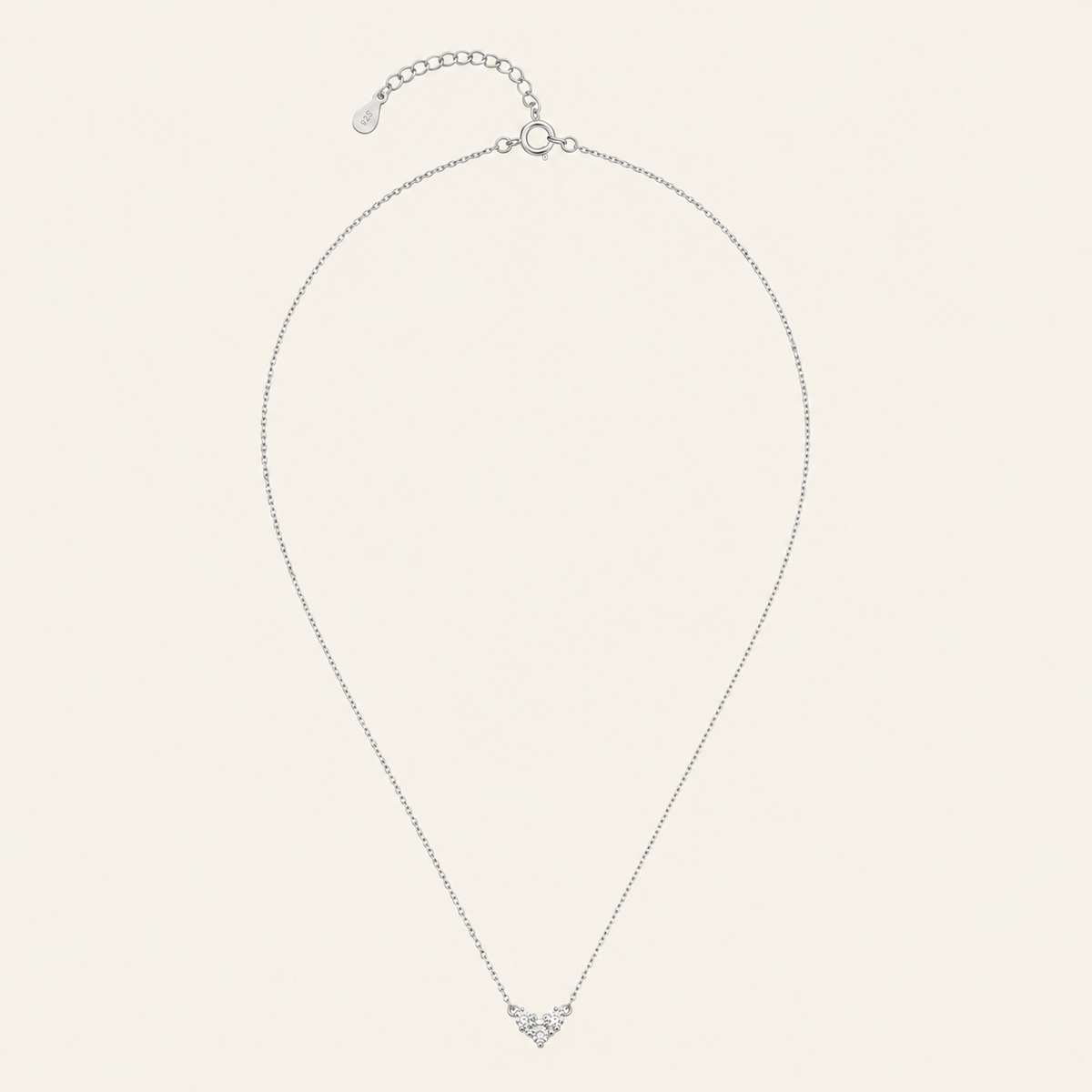 A delicate silver chain necklace with a small heart-shaped pendant set with clear stones, displayed on a light cream background.