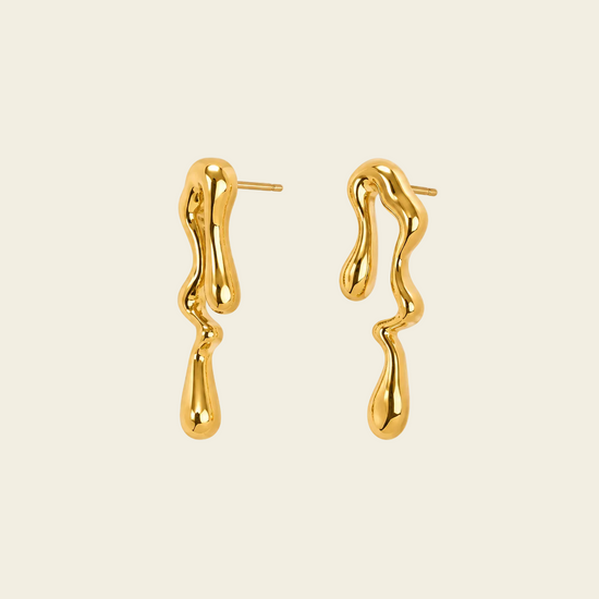 A pair of gold dangling earrings with a dripping, organic wavy shape, shown on a solid pale beige background.