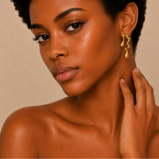 A close-up portrait of a woman with short dark hair wearing a single abstract, wavy gold dangle earring against a warm beige background.