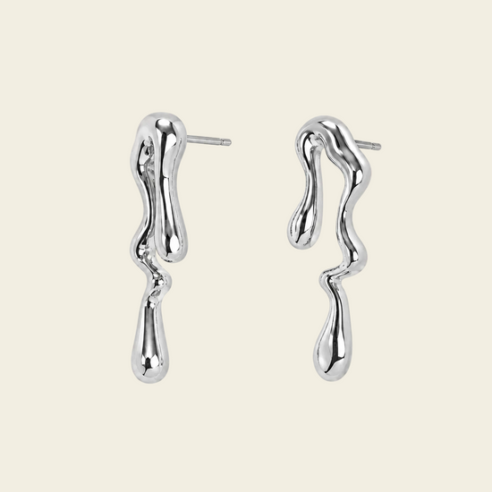 A pair of polished silver drop earrings with a wavy, dripping organic shape, displayed on a solid pale peach background.