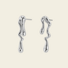A pair of polished silver drop earrings with a wavy, dripping organic shape, displayed on a solid pale peach background.