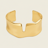 A polished silver cuff bracelet with a wide, sculptural, wavy profile and a central V-shaped notch, shown on a solid light beige background.