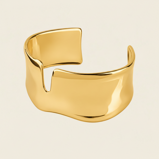 A polished gold cuff bracelet with a unique wavy, open design, shown on a solid light beige background.
