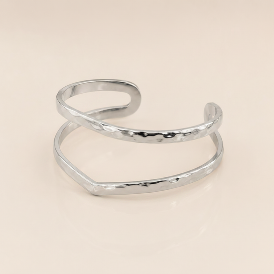 A silver-colored open cuff bangle with a hammered texture, displayed on a clean, light beige background.