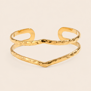 A gold-tone open cuff bangle bracelet with a hammered texture, displayed on a light cream background.