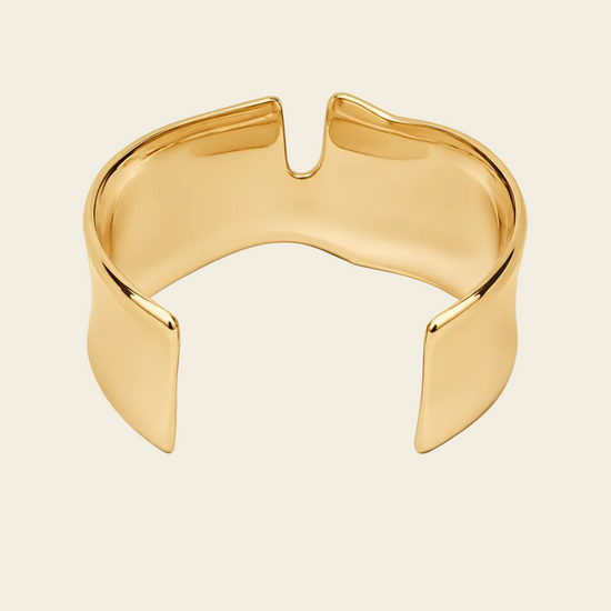 A polished gold open cuff bracelet with a unique, organic wavy shape, shown on a light cream background.