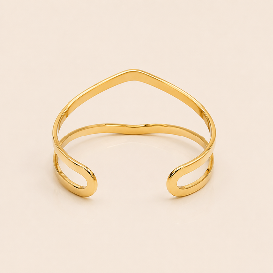A polished gold open cuff bangle with a wavy, organic line design, photographed on a solid light beige background.