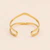 A polished gold open cuff bangle with a wavy, organic line design, photographed on a solid light beige background.