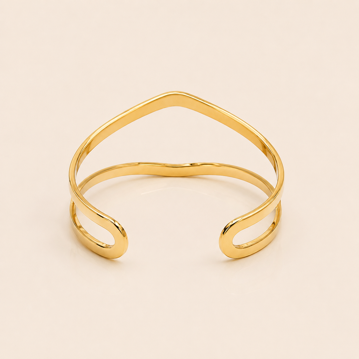 A polished gold open cuff bangle with a wavy, organic line design, photographed on a solid light beige background.
