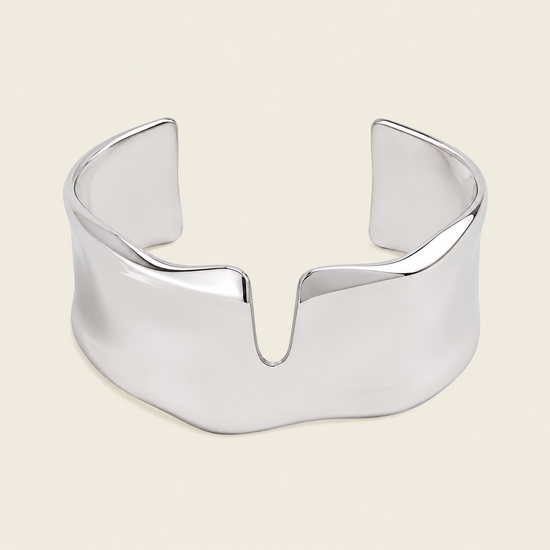 A polished silver cuff bracelet with a wide, sculptural, wavy profile and a central V-shaped notch, shown on a solid light beige background.