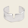 A polished silver cuff bracelet with a wide, sculptural, wavy profile and a central V-shaped notch, shown on a solid light beige background.