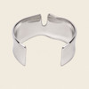 A polished silver-toned metal cuff bracelet with a unique curved, open design, shown on a solid light beige background.