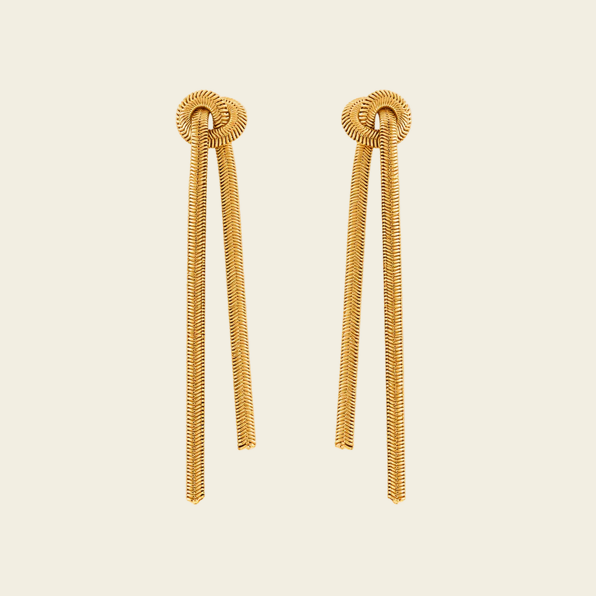 A pair of gold-toned statement earrings with textured circular studs and long, snake-like draped tails, shown on a solid light beige background.