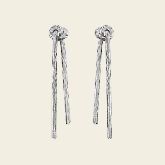 A pair of silver-toned dangle earrings with textured circular studs and long, snake-like draped chains, shown on a plain light beige background.