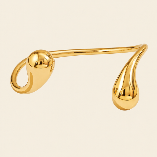 A polished gold-toned metal cuff bracelet with a curved, organic shape featuring a rounded loop on one end and a teardrop-shaped end on the other, set against a solid light beige background.