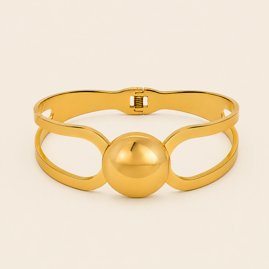 A polished gold-tone metal cuff bracelet with a central spherical bead and openwork loop details, shown on a light cream background.