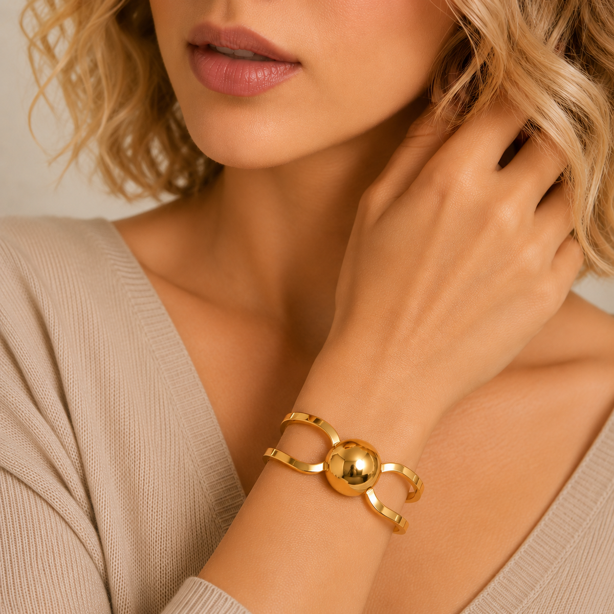 A close-up of a woman wearing a polished gold wrap bracelet with a central spherical bead, styled with a beige ribbed knit top.