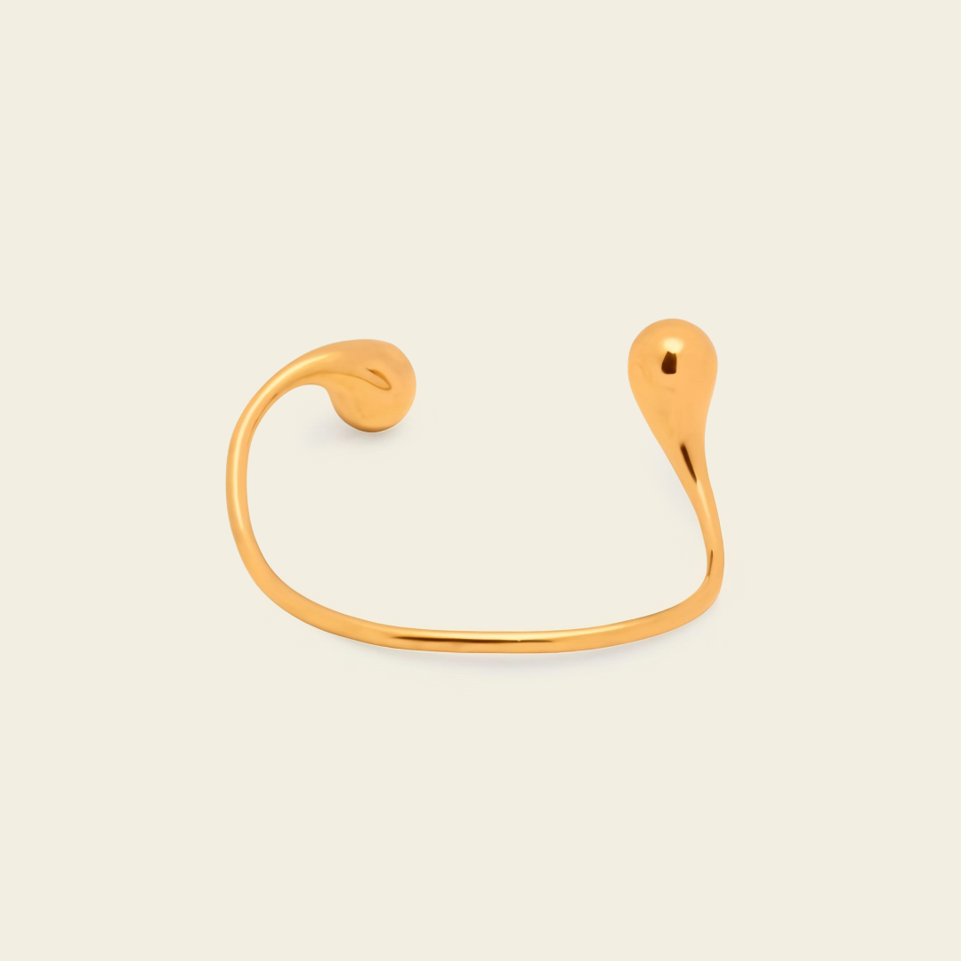 A minimalist gold open cuff bracelet with a curved, organic teardrop-shaped end, shown on a light cream background.