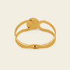 A polished gold-toned metal cuff bracelet with a unique openwork design and a small sliding clasp, shown on a solid light beige background.