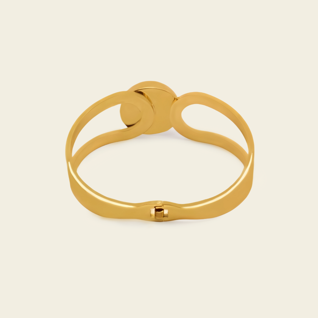 A polished gold-toned metal cuff bracelet with a unique openwork design and a small sliding clasp, shown on a solid light beige background.