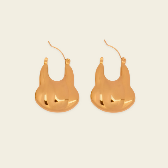 A pair of gold-toned, abstract sculptural hoop earrings on a light cream background.
